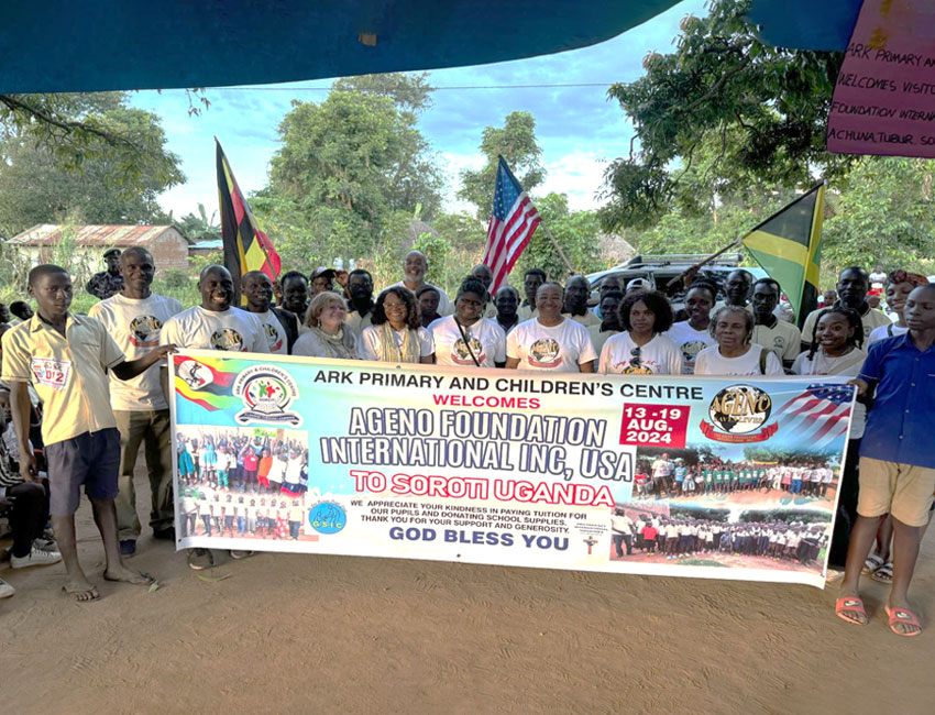 A diverse group of people stand outdoors holding a large banner for the "Ageno Foundation International Inc, USA" event in Soroti, Uganda, dated August 13-19, 2024. Flags and trees can be seen in the background.