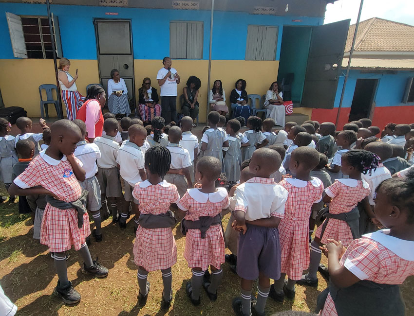 A group of young students in uniforms, with checkered dresses for girls and white shirts with gray shorts for boys, are gathered outdoors. They are facing adults, some seated and some standing, addressing them in front of a colorful building.