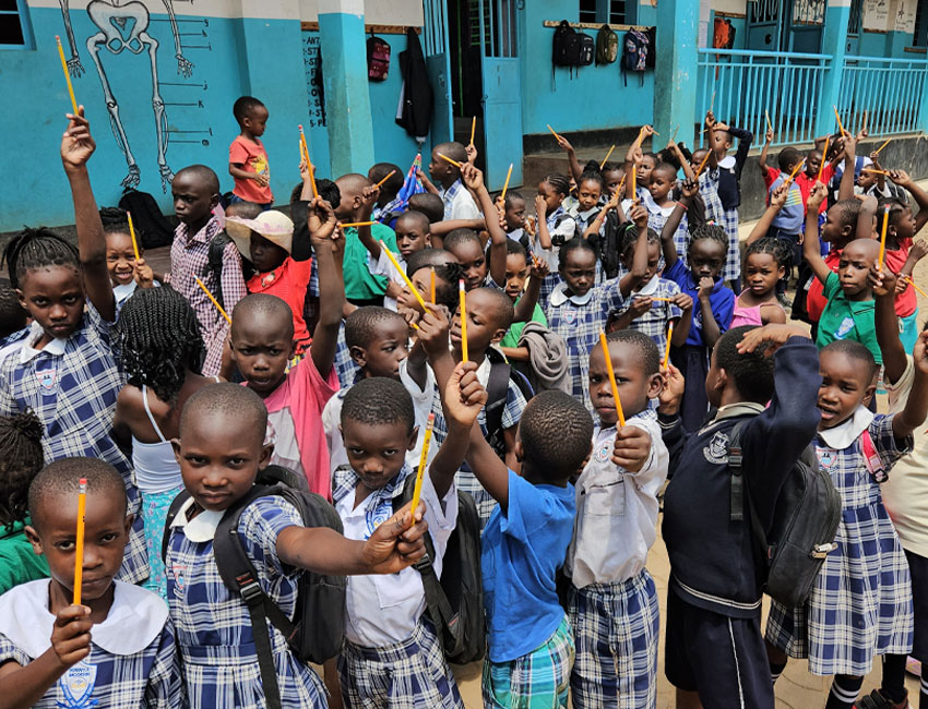 A group of schoolchildren in plaid uniforms hold up pencils enthusiastically. They are gathered outside, standing closely together, with a blue building in the background.