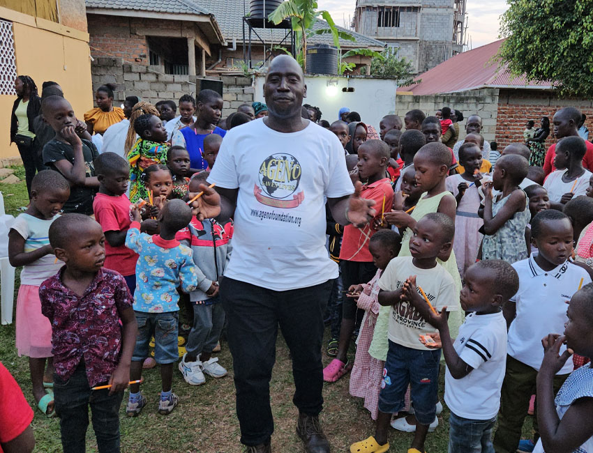A man in a white t-shirt stands smiling among a group of children outdoors. The children, of various ages, gather around him. Some people are standing in the background near buildings and vegetation.