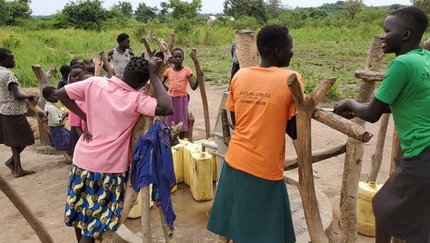 A group of people, mostly children, gather around a communal water well in a rural area. They use hand pumps made from wood and metal to fill yellow jerry cans. Lush green vegetation and trees are visible in the background, indicating a tropical environment. Your charity donation can help bring clean water to more villages like this one.