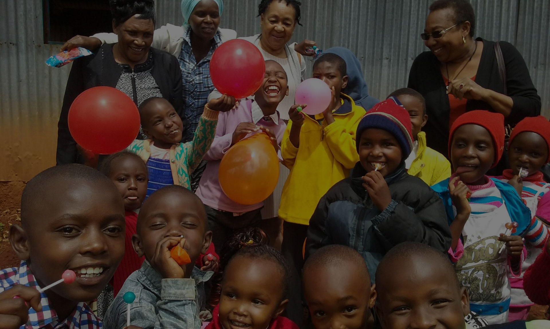 A joyful group of children and adults gather in front of a corrugated metal wall. The children, clutching colorful balloons and lollipops, smile and laugh together. Even the local veterinarian joins in the merriment with the adults in the background, creating a vibrant and happy scene.