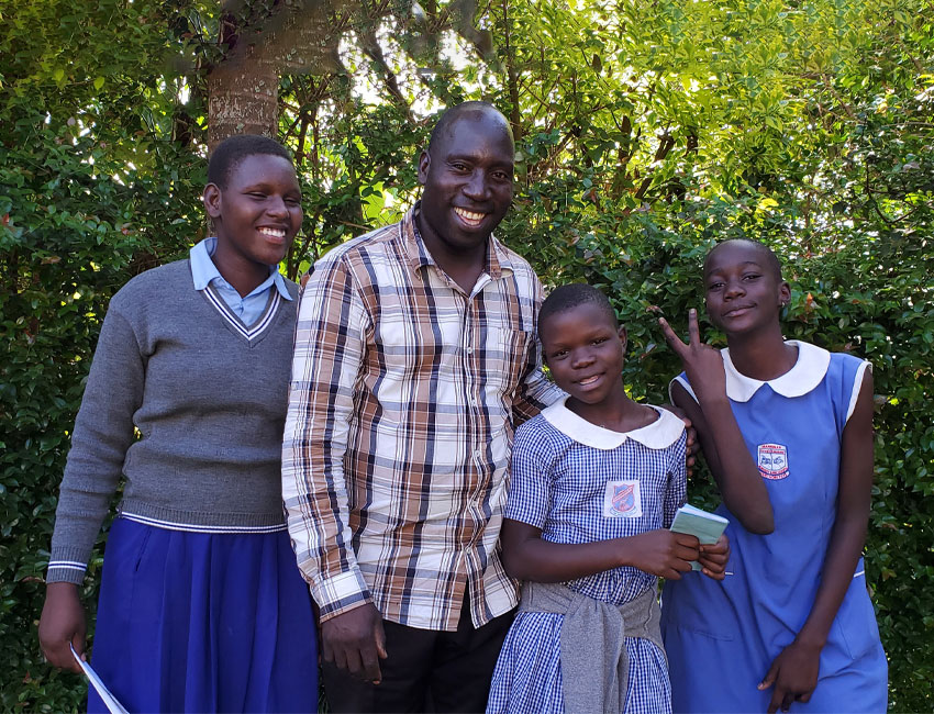 A group of four people standing outdoors in front of leafy greenery. From left to right, two school girls in uniforms, a man in a plaid shirt, and another school girl in uniform are all smiling; one girl on the right holds up a peace sign, celebrating their recent charity water donation.
