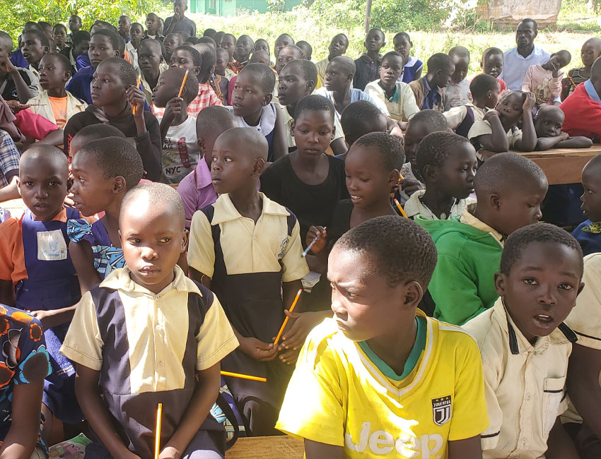 A large group of school children in uniforms sits together outdoors. Some hold pencils and notebooks, attentively focused on something ahead. The background shows a green, leafy environment with a few adults standing among the children, possibly organizing charity donations or teaching about water conservation.