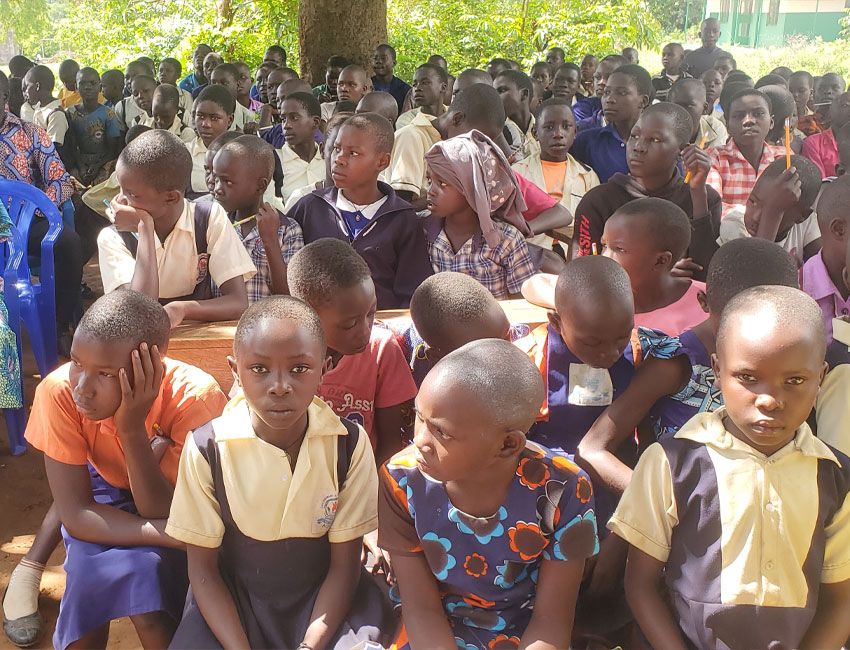 A group of young students seated closely together on wooden benches outdoors. Many of the children are wearing school uniforms, and some are attentively looking forward while others seem distracted. A tree and greenery can be seen in the background. Nearby, a water station set up through local charity donations supports their daily needs.
