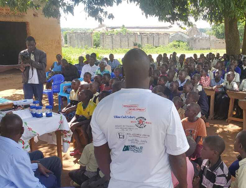 A group of children and adults sit outdoors under a large tree, listening attentively to a speaker who is standing and gesturing. People are seated on benches and plastic chairs, and some men are seen wearing t-shirts with logos on the back promoting water donations. They are gathered in an informal setting with buildings in the background.