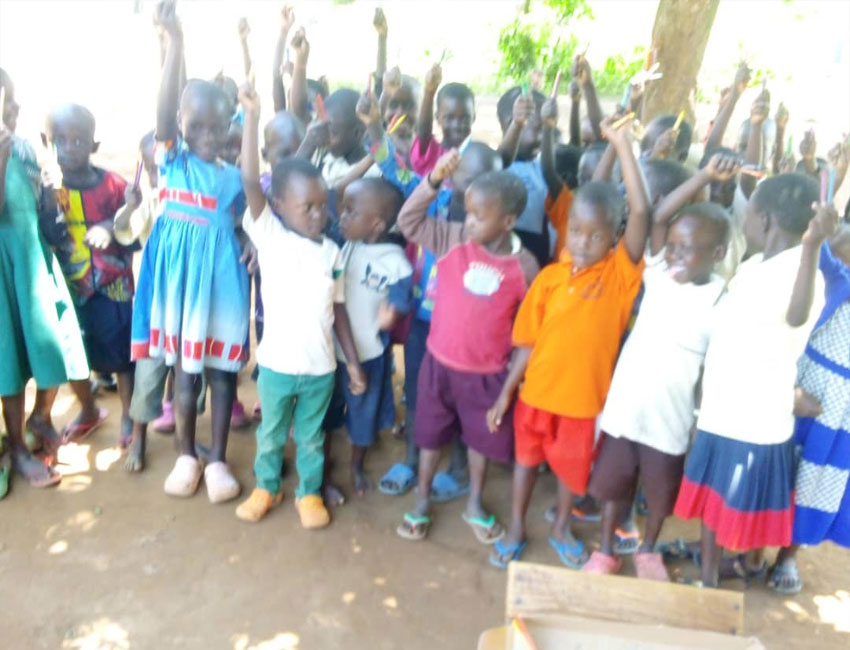 A group of young children stand outdoors, some with their arms raised enthusiastically. They are dressed in various colorful clothes and appear joyful and engaged. Trees and natural light are visible in the background, suggesting a sunny day. This cheerful scene was made possible through a water charity donation.