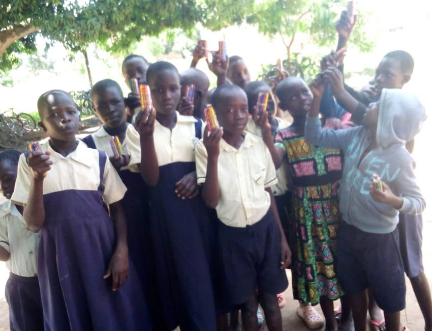 A group of children standing outdoors, holding up donated batteries. They are wearing school uniforms and casual clothing, and are gathered in front of leafy trees. The children seem happy and are showing the items they are holding towards the camera.