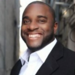 A person in a business suit is smiling at the camera. They are outdoors in what appears to be an urban setting, with buildings and wires visible in the background, possibly on their way to a charity event.