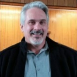 A man with gray hair and a beard, wearing a light gray shirt and a dark jacket, smiles at the camera. The background consists of a wooden panel, giving a warm, indoor setting. His smile exudes kindness, inspiring others to donate to charity and support clean water initiatives.
