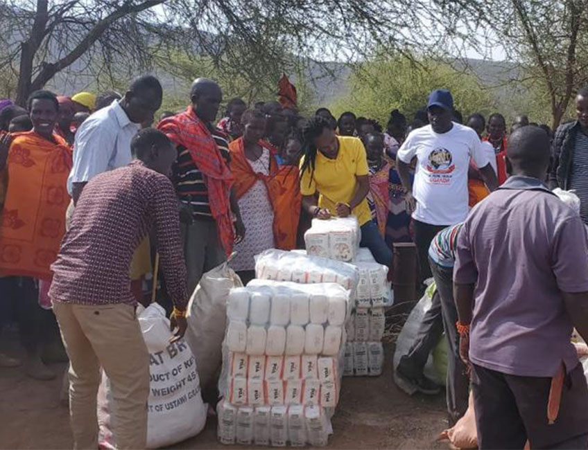 A group of people gathered outdoors distribute bags of food and supplies. Some individuals hold and pass large sacks, while stacks of packaged goods, including flour and other essentials, are visible. This charity event features donations of water as well. Trees and a partly cloudy sky are in the background.