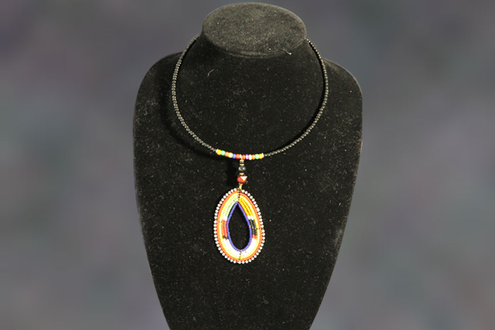 A black beaded necklace with a large, multi-colored teardrop pendant displayed on a black stand. The pendant features concentric patterns of yellow, white, orange, and blue beads, with a small red bead at the top. A portion of sales will go towards charity efforts to provide clean water to those in need.