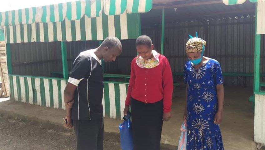 Three people are standing in front of a green and white striped building. The man is wearing a black and white shirt, while the two women are dressed in colorful outfits, one in red and the other in blue. One woman is holding a bag that says "Donate," and they appear to be looking down.