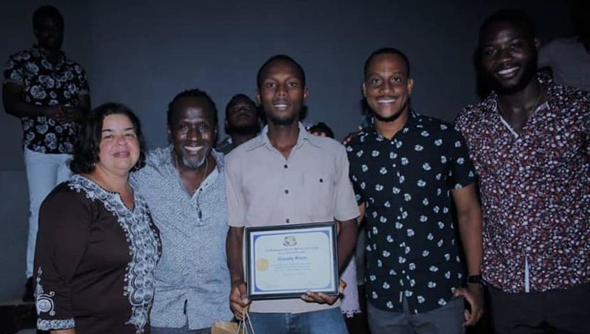 A group of five people stand together smiling. The person in the center holds a certificate, likely from a recent charity event. They are in a dimly lit room, and one person is slightly in the background, mostly obscured. The people are casually dressed.
