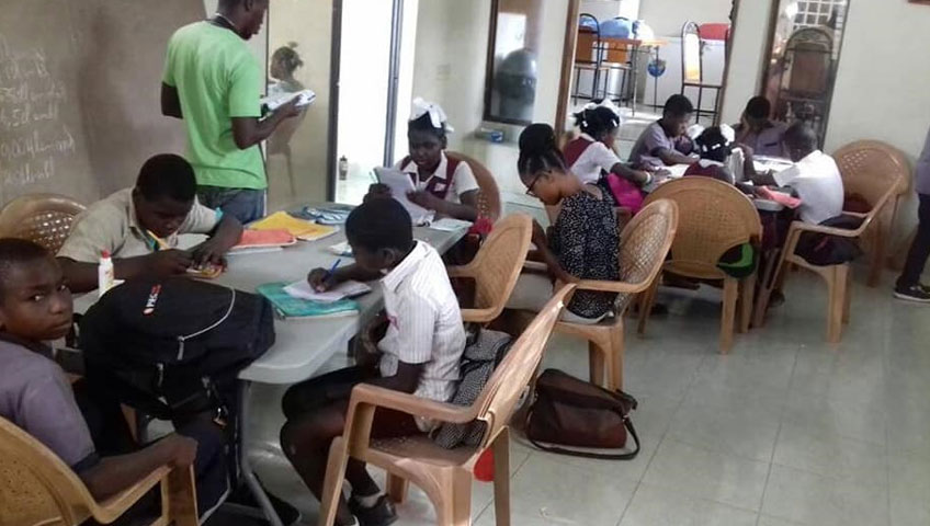 A classroom scene with several students seated around tables, focused on writing or reading. A person stands next to the blackboard, holding papers that explain an upcoming charity event. The room is equipped with chairs, tables, and some visible classroom supplies, including a donation box for clean water projects.