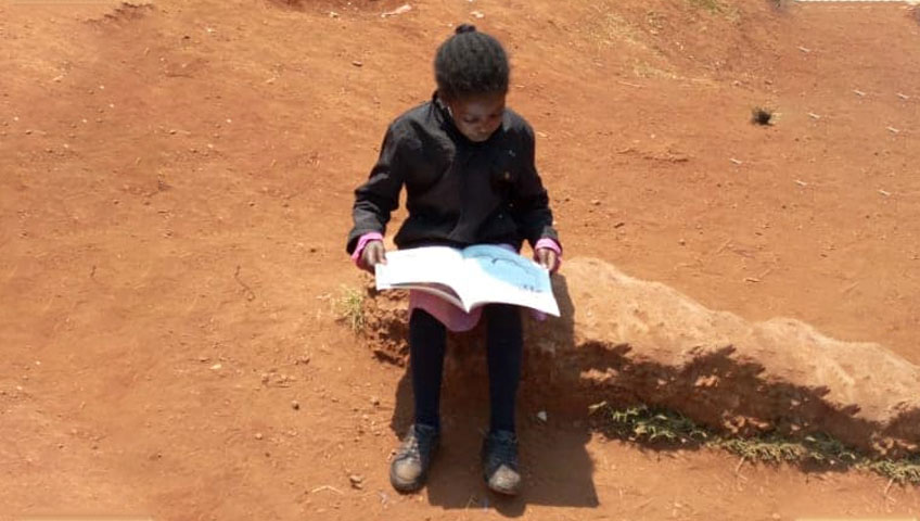 A child sits on a patch of bare, reddish ground while reading a large book, fully absorbed in its contents. The child wears a dark outfit and appears to be alone in the outdoor setting, surrounded by an arid landscape where clean water is scarce—a stark reminder of the need for charity and donation.