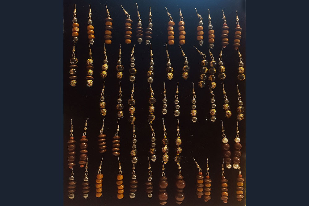 A collection of handmade earrings displayed in rows against a dark background. Each pair features a series of stacked round beads in varying colors and patterns, including brown, gold, and silver. The earrings hang from simple hooks. Proceeds from sales will be donated to water charity initiatives.