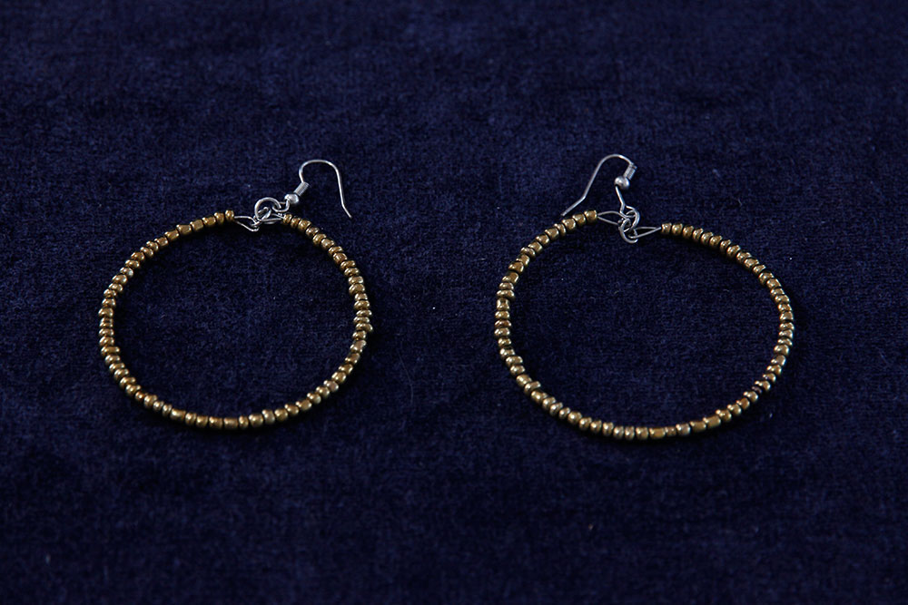 A pair of gold beaded hoop earrings with simple hooks set against a dark textured background. The beads are uniform in size, forming a circular shape for each earring, perfect for adding a touch of elegance while supporting charity through your purchase.