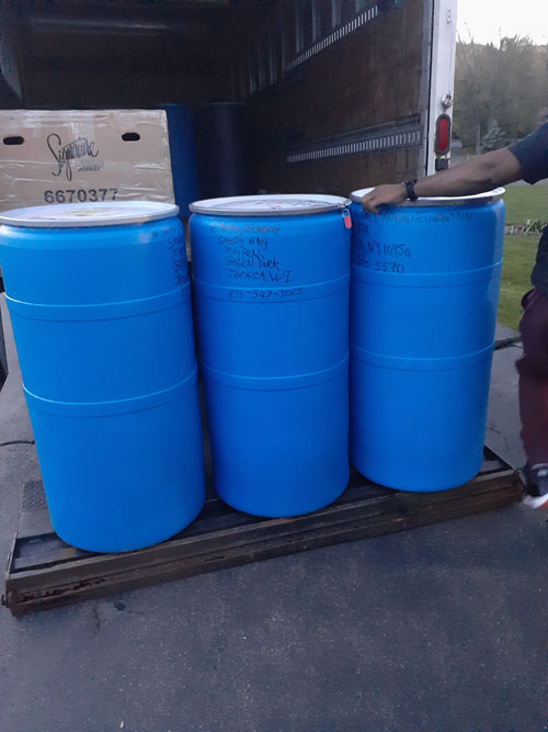 Three large blue plastic barrels, likely containing donated water, stand side by side on a wooden pallet inside a truck. The barrels have handwritten labels on the white lids. The truck's rear door is open, revealing a partially visible person on the right.