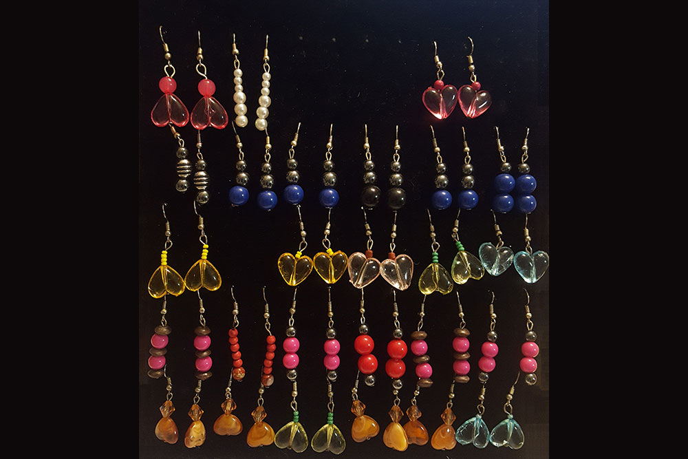 A display of various dangle earrings arranged in rows on a black background. Designs include heart-shaped charms, pearl beads, and colorful round beads in hues of red, pink, blue, yellow, and green. Each unique pair supports a water donation initiative with every purchase.