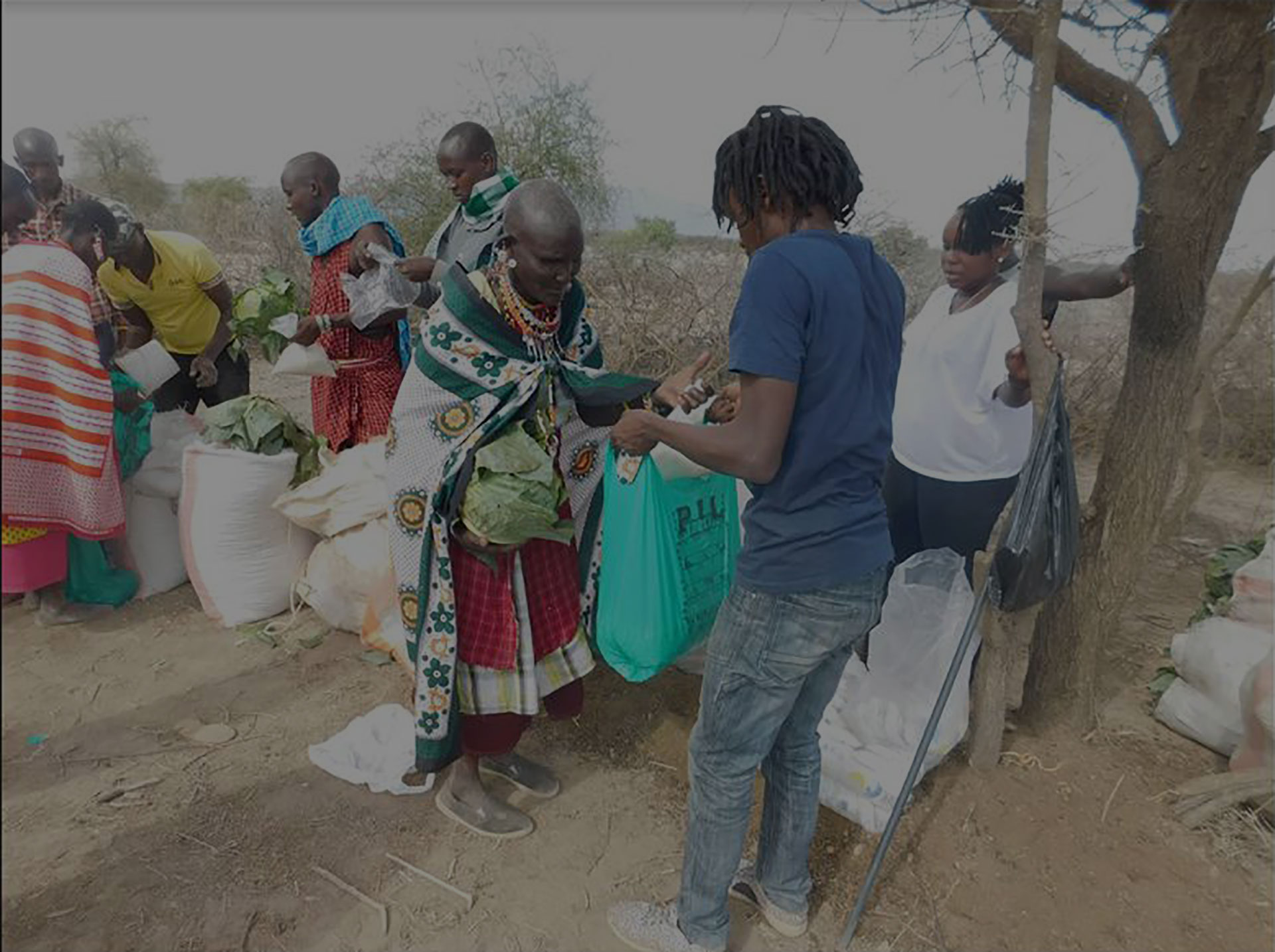 A group of people engage in a charitable distribution of goods in an outdoor setting. One person hands a green bag to another, who wears traditional attire. Several others participate in the background, with bags and items around them. A tree is visible to the right.