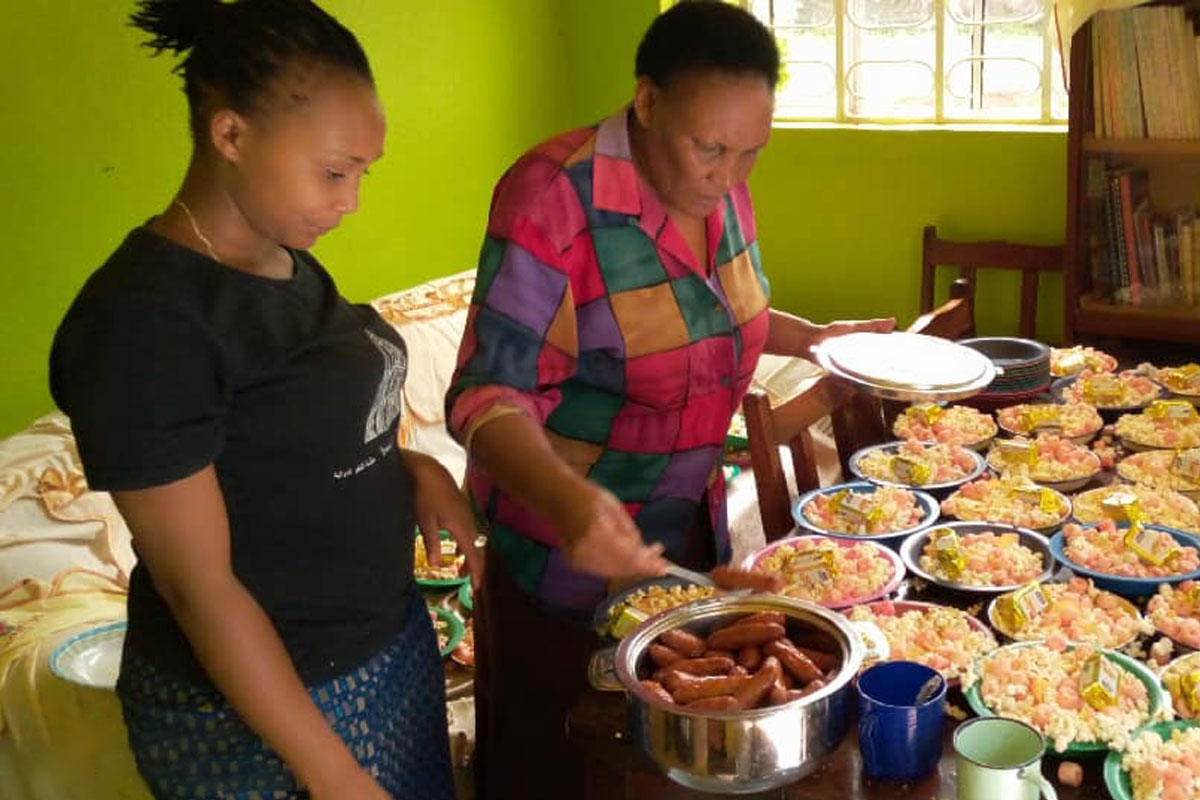 Two women are preparing food in a room with green walls. The table is filled with plates of food, including sausages, rice, and vegetables. One woman, a veterinarian in a colorful blouse, holds a plate while the other, in a black t-shirt, stands beside her.