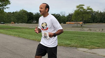 A man is jogging on a paved path in a park-like setting, participating in a charity race. He is wearing a white T-shirt with a graphic on the front, black shorts, and a race bib numbered 247. Trees and grassy areas are in the background, and he carries a bottle of water to stay hydrated.