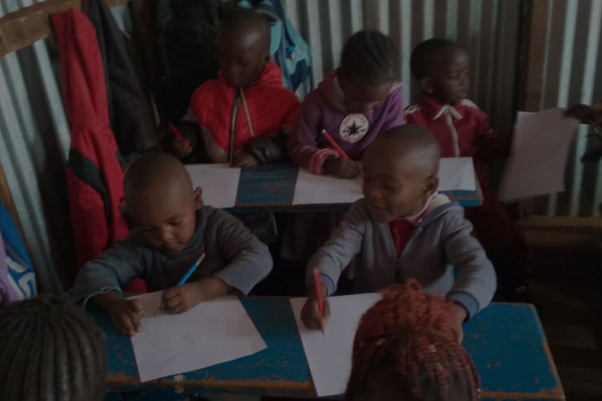 A group of children sitting at blue desks inside a classroom, focused on drawing or writing on sheets of white paper. The room has corrugated metal walls, and the children are wearing hoodies and jackets. The lively environment showcases the impact of charitable donations as they engage in their activities.