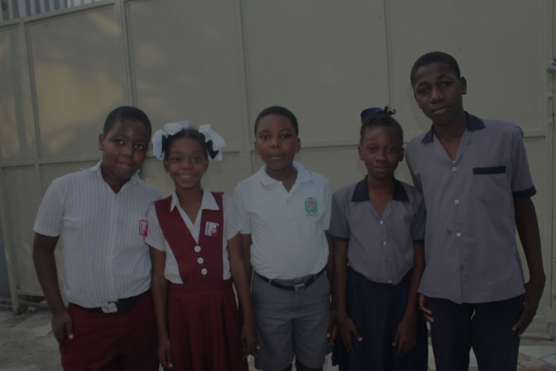 A group of five children stand together, smiling in front of a beige wall. Dressed in various school uniforms—some in shirts and skirts or shorts, others in dresses—they pose happily for the photo. Their bright smiles radiate a sense of community, perhaps thanks to a recent charity donation that brought clean water to their school.