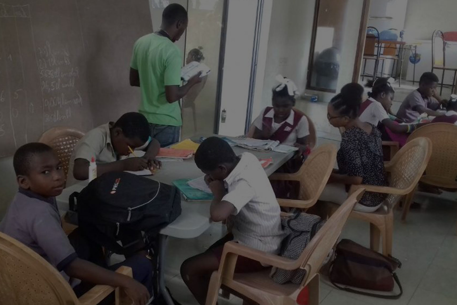 Several children and an adult are gathered inside a classroom. Some children are seated at desks working on tasks, while others are standing or moving around. The adult is looking at a book, and there are chalkboard writings and various classroom materials visible, encouraging students to donate water to those in need.