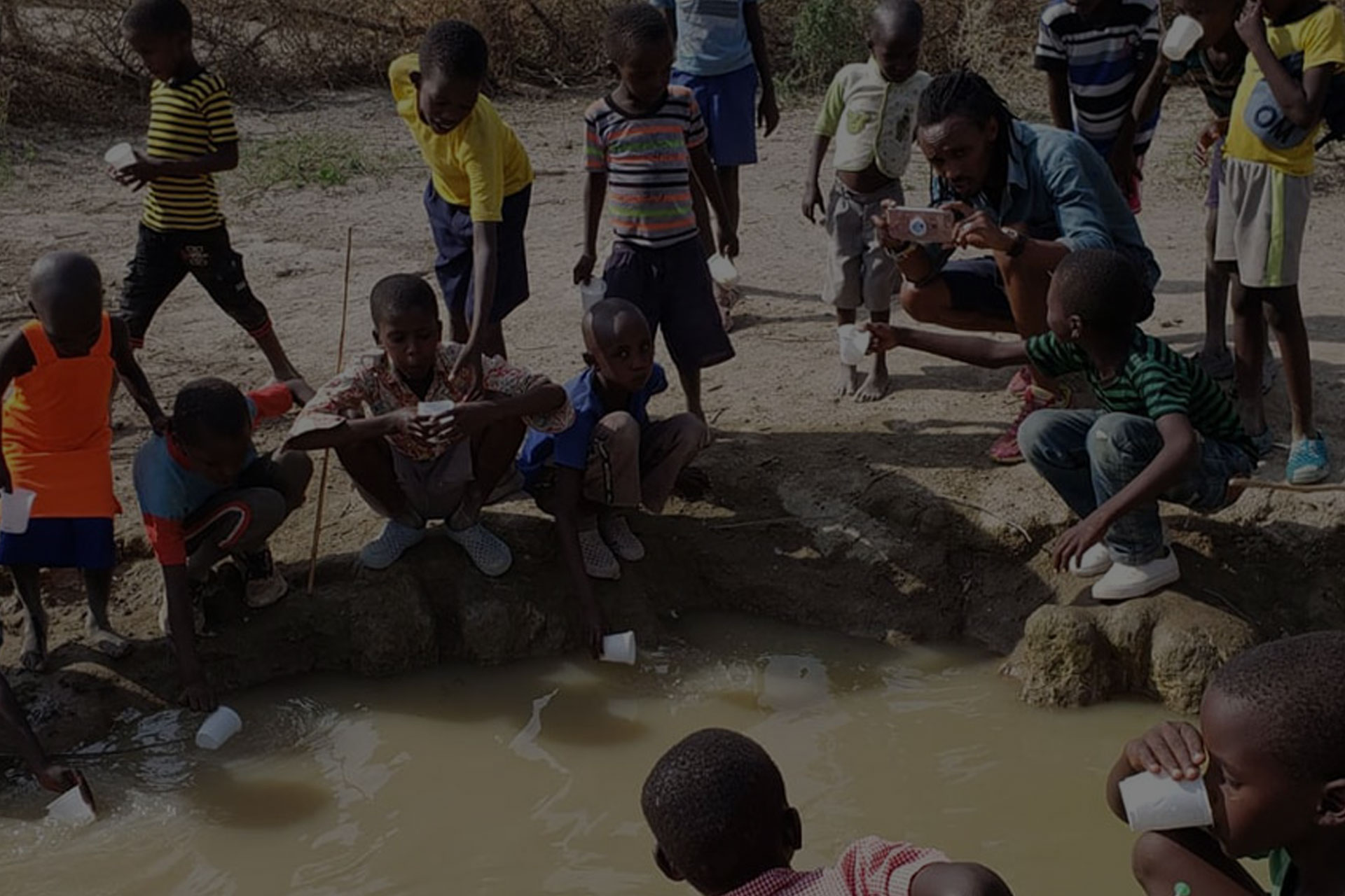A group of children gather around a small, muddy body of water. Some are holding containers, seemingly collecting water. An adult crouches down, holding a camera, capturing the scene for a charity dedicated to providing clean water to rural communities.