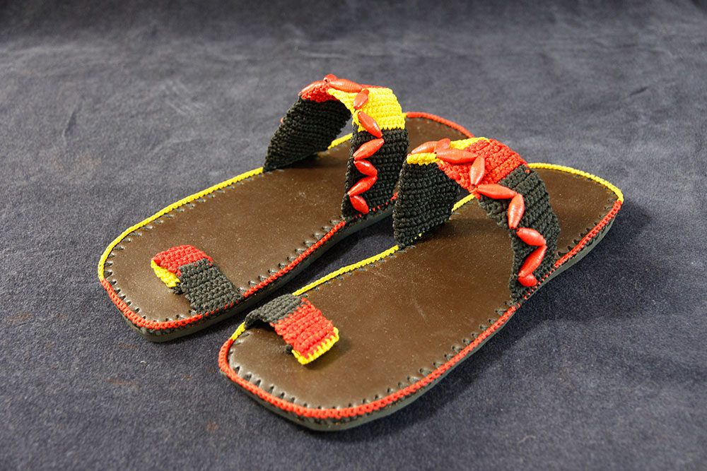 A pair of handmade sandals with a brown leather sole and colorful straps rests on a dark blue fabric background. The woven pattern in yellow, red, and black alternates in a zigzag design. A portion of each purchase is donated to provide clean water to communities in need.